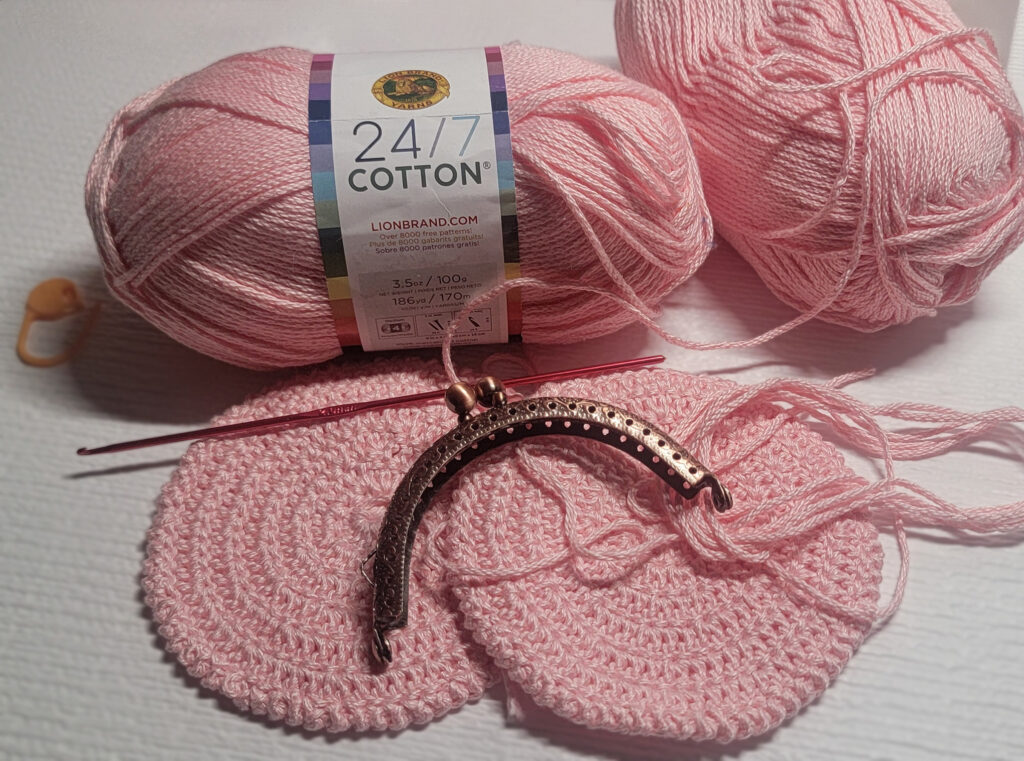 This image showcases the materials used for a spring-themed crochet rose clasp purse project, emphasizing the vibrant pastel pink yarn and crafting tools. Two skeins of Lion Brand 24/7 Cotton yarn in the Pink Lemonade colorway, a crochet hook, a stitch marker, and two small, crocheted circles displayed on a white background. 